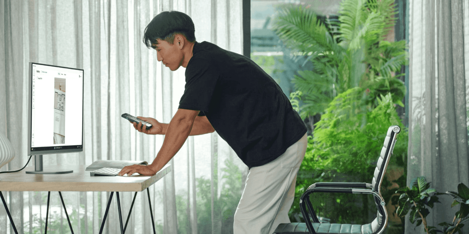 Hombre trabajando en escritorio de pie con computadora y teléfono inteligente en oficina en casa moderna con plantas